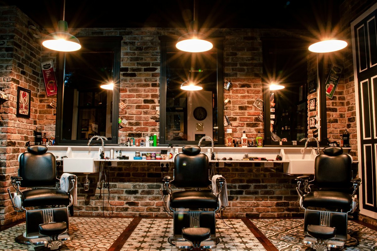 Three barber chairs facing mirrors along a brick wall under hanging lamps
